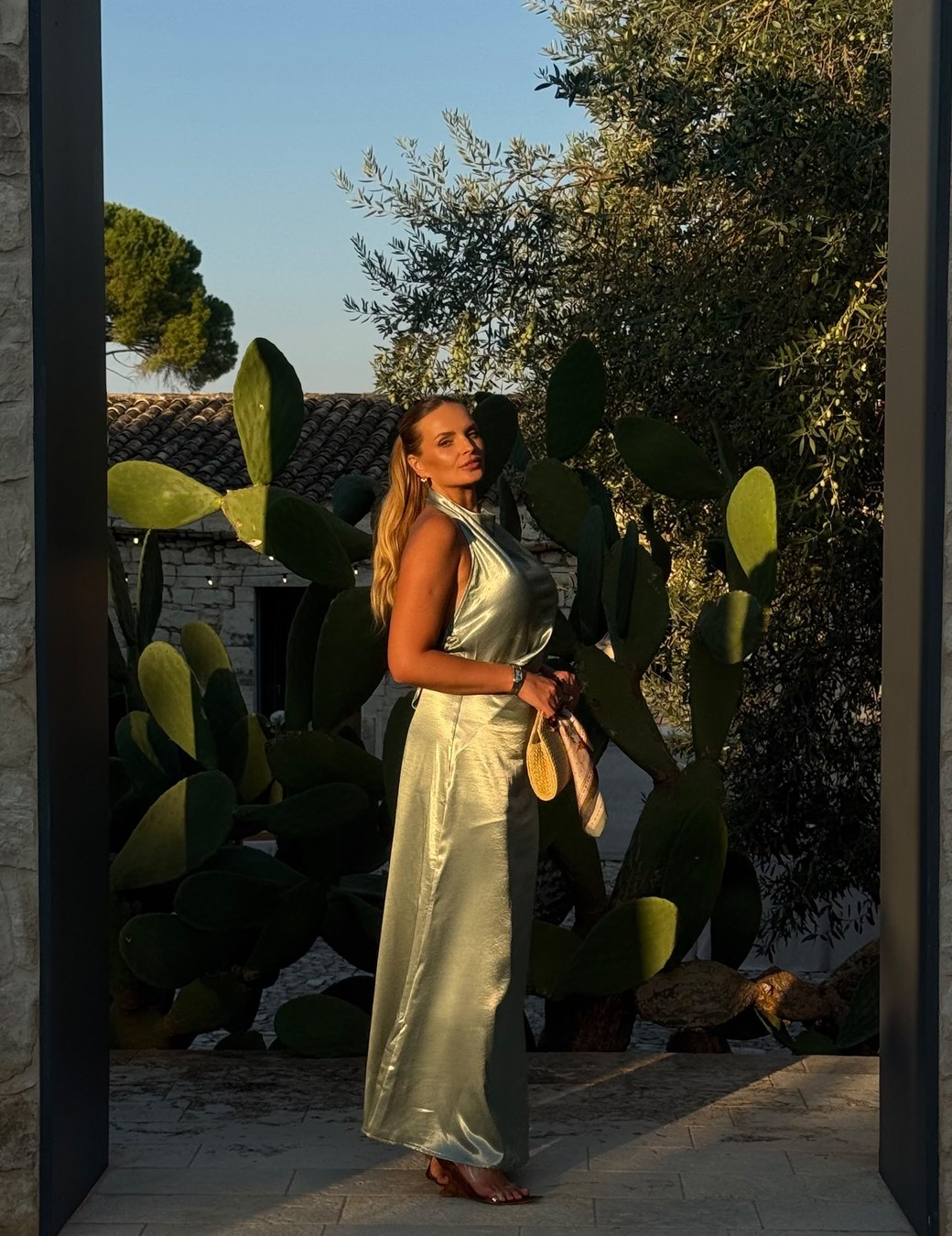 Joice in a long satin dress, turned to the side and centred inside a stone doorway, with cacti and an olive tree behind her at golden hour