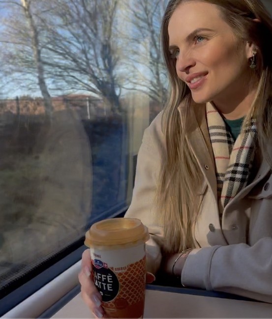 Joice smiling by a train window, holding a coffee cup